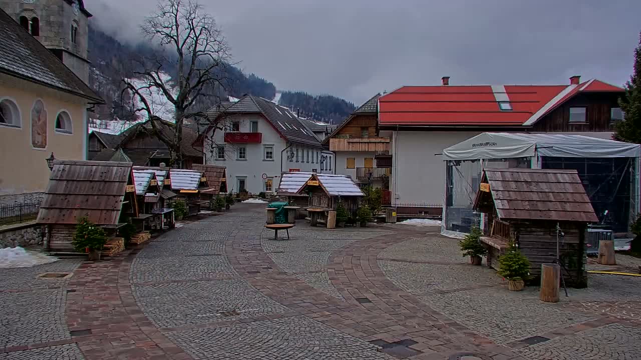 A cobblestone town square features numerous rustic wooden stalls and traditional buildings against a backdrop of snow-dusted mountains, all under a grey and overcast sky.