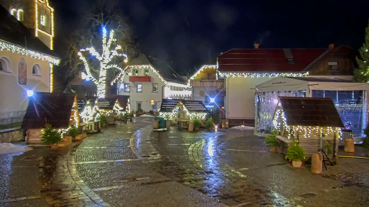 A cobblestone square is festively illuminated at night with numerous string lights adorning buildings, a large tree, and market stalls, their reflections shimmering on the wet ground under a dark, wintry sky with patches of snow.