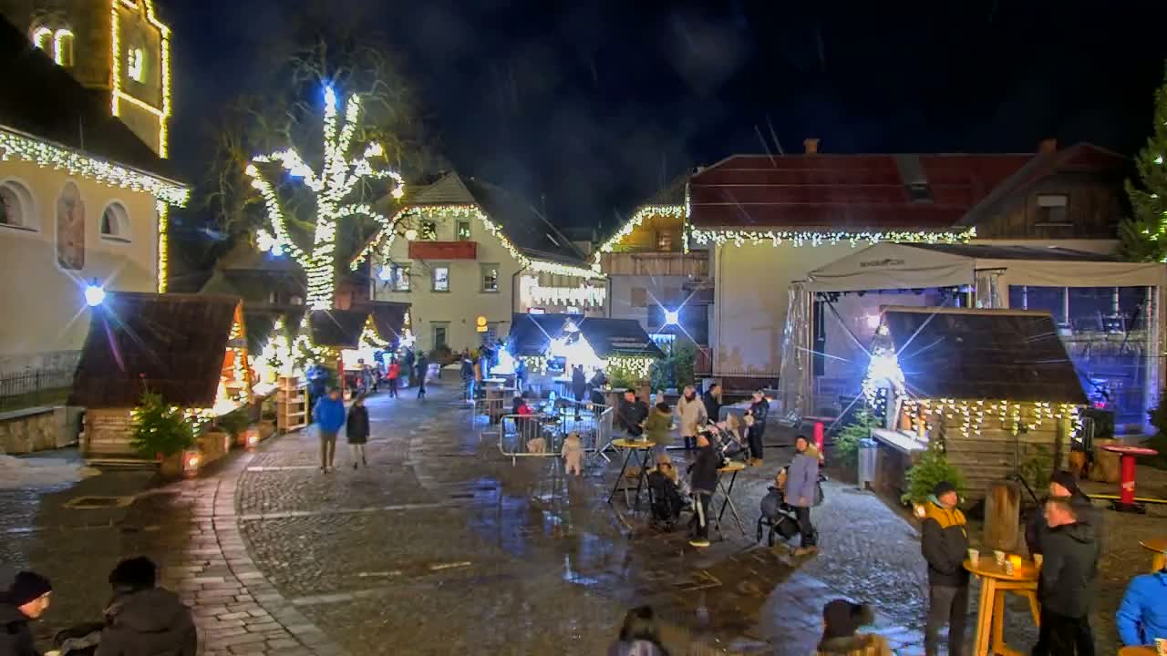 A lively Christmas market is seen at night, with people gathered around wooden stalls and tables, amidst buildings and a large tree sparkling with festive lights, all on wet cobblestone ground under a clear dark sky.