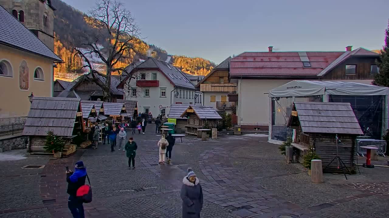 A lively Christmas market is seen at night, with people gathered around wooden stalls and tables, amidst buildings and a large tree sparkling with festive lights, all on wet cobblestone ground under a clear dark sky.