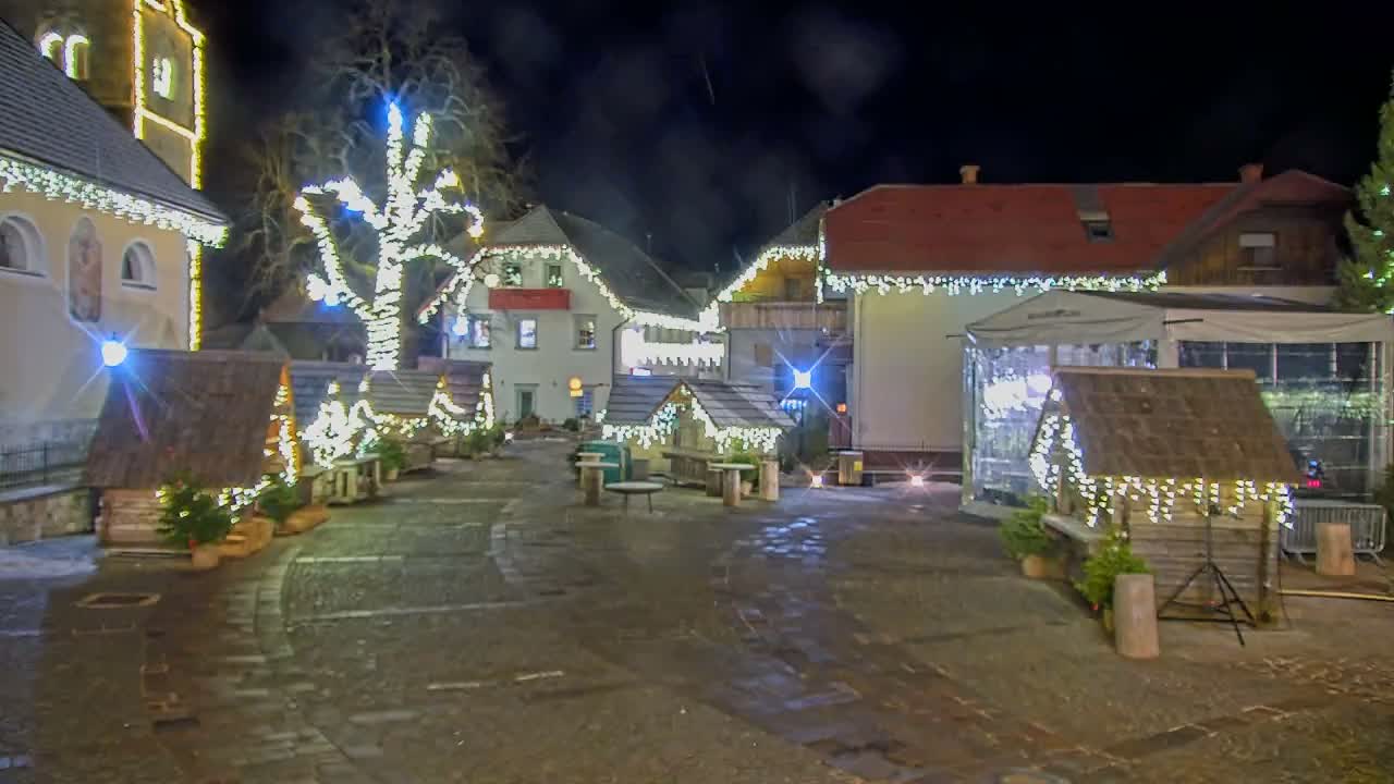 A lively Christmas market is seen at night, with people gathered around wooden stalls and tables, amidst buildings and a large tree sparkling with festive lights, all on wet cobblestone ground under a clear dark sky.