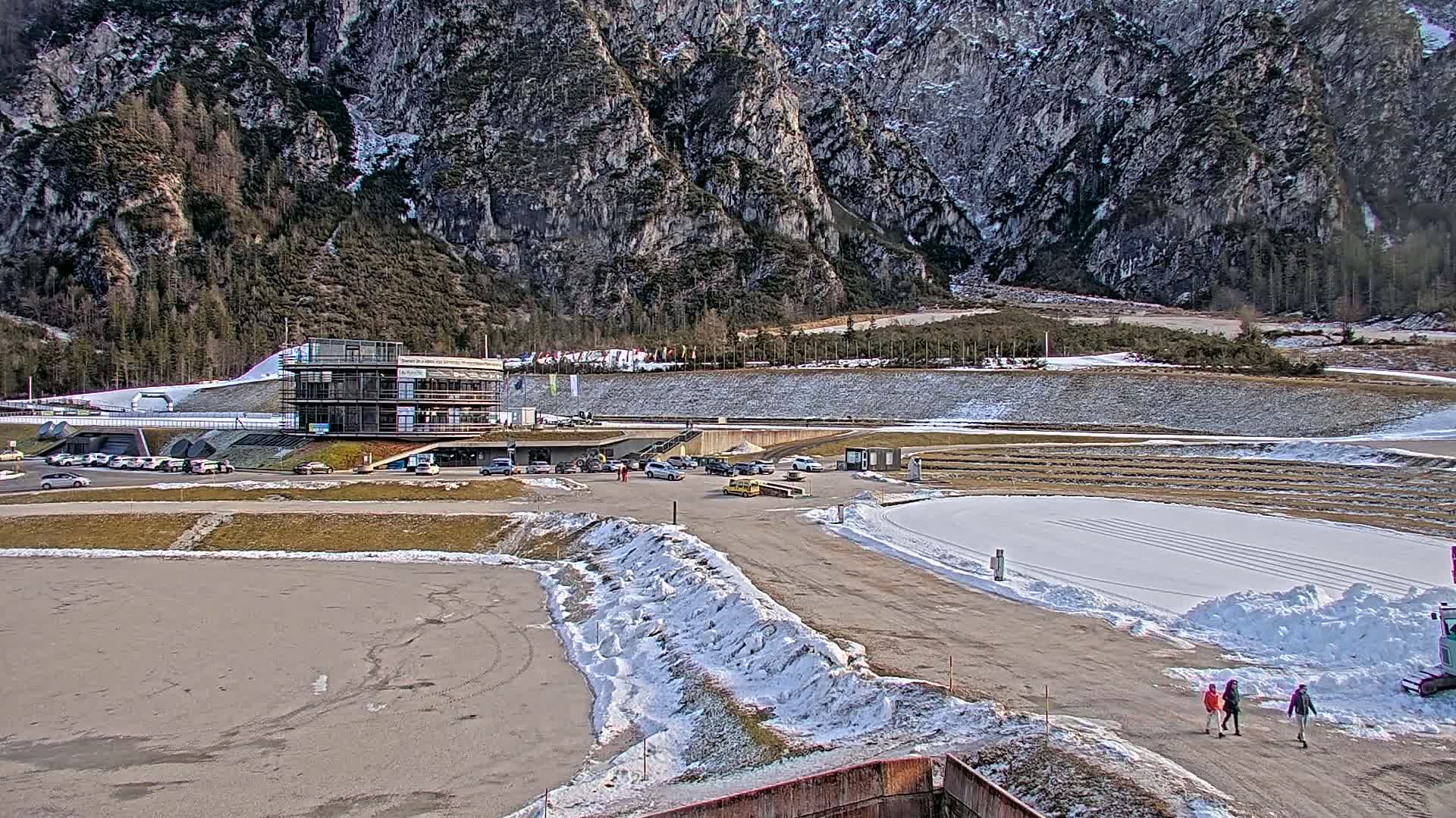 A modern building sits near a snowy track at the foot of a large, rocky mountain range.