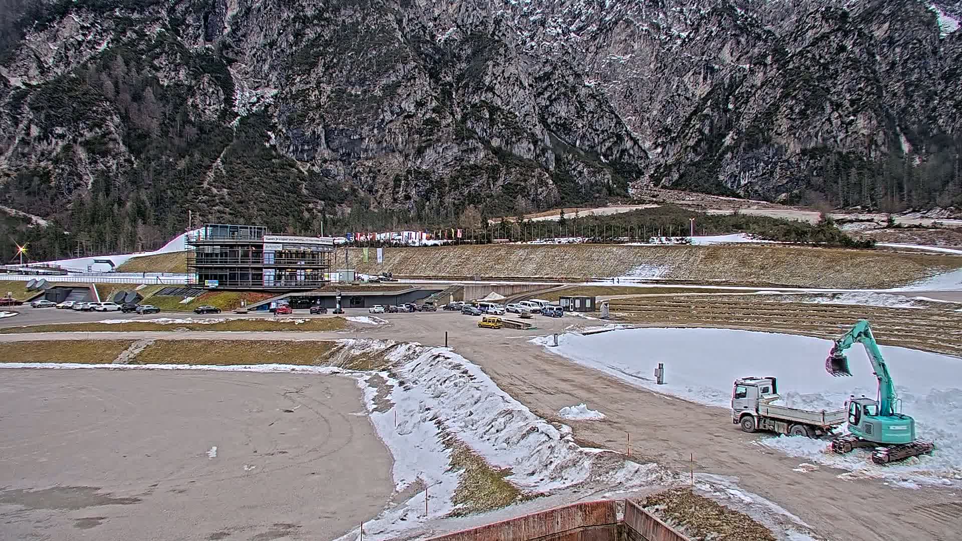 A snow-dusted parking lot and a building complex sit at the base of a large, rocky mountain under an overcast sky, with a small excavator and truck working nearby.
