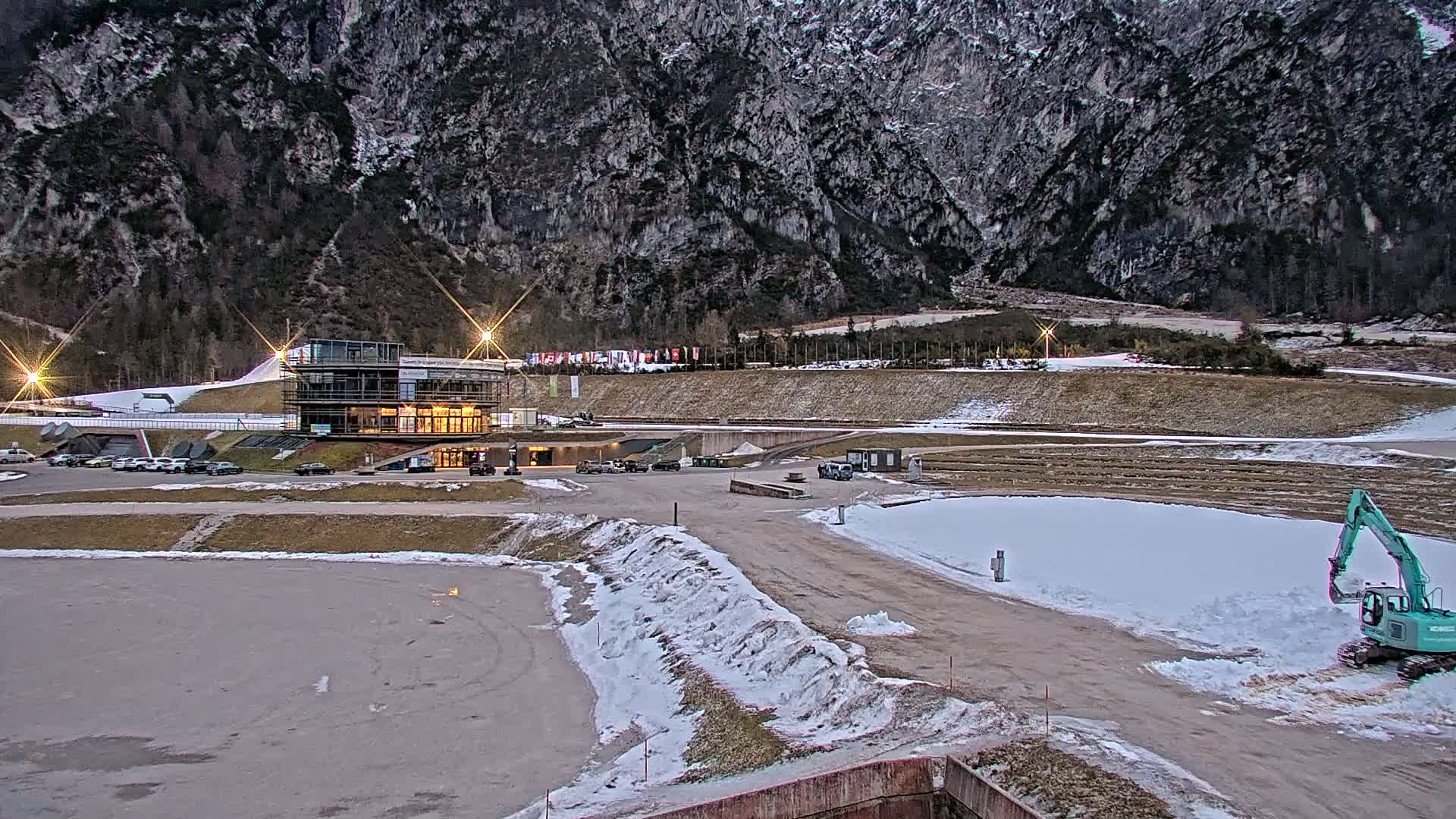 A modern building sits at the base of a large, dark mountain range in a snowy, outdoor setting, with a small excavator working nearby.