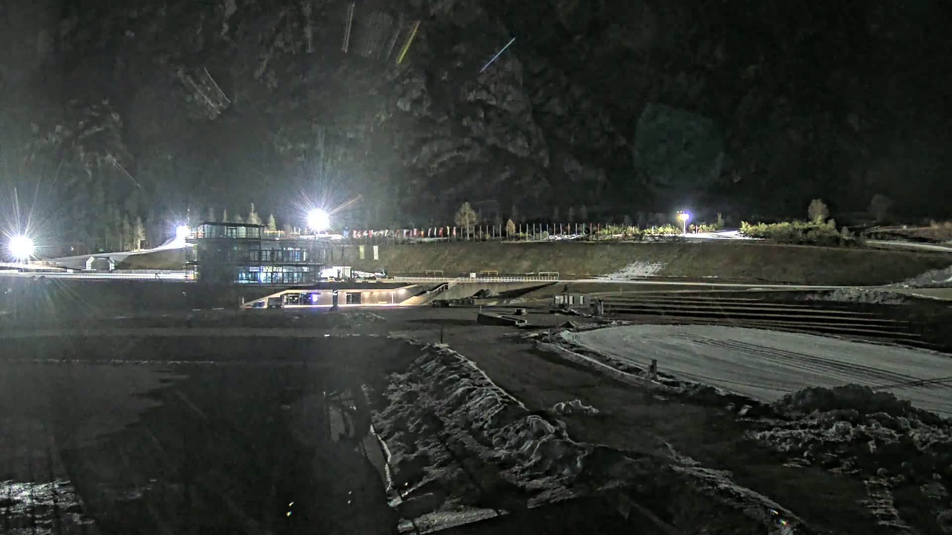 A nighttime view of a snow-covered outdoor sports facility with a multi-story building, illuminated by bright lights.