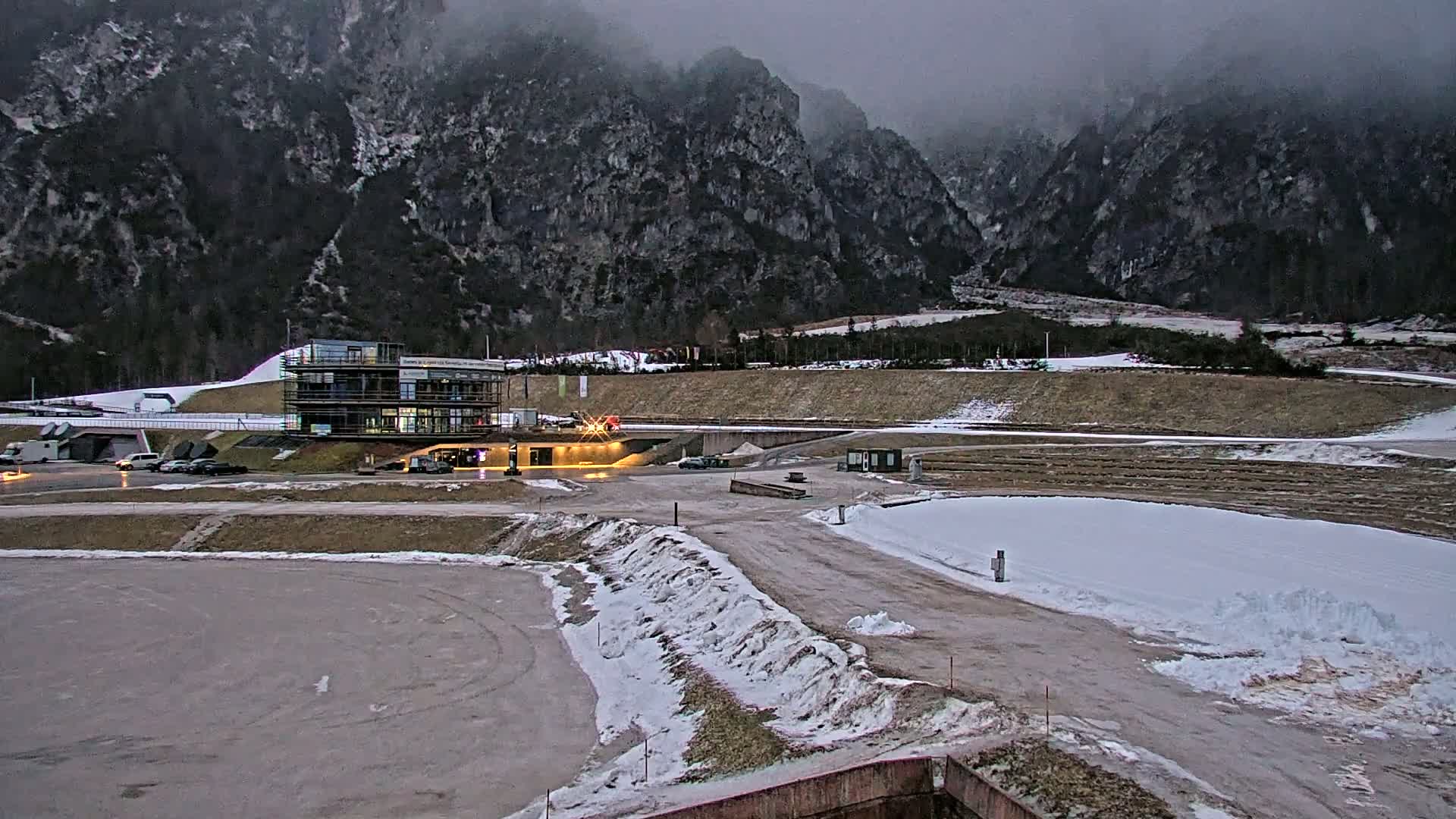 A multi-story building under construction sits in a snowy parking lot near a mountain range under an overcast sky.