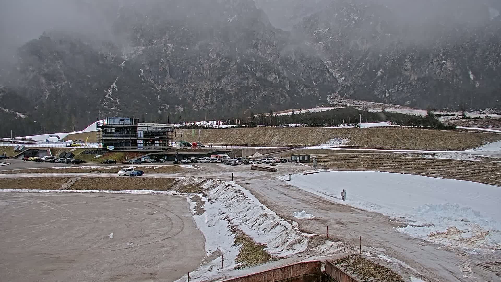 A parking lot with several cars sits in front of a multi-story building under a cloudy sky, with snow partially covering the ground and a mountainous background shrouded in mist.
