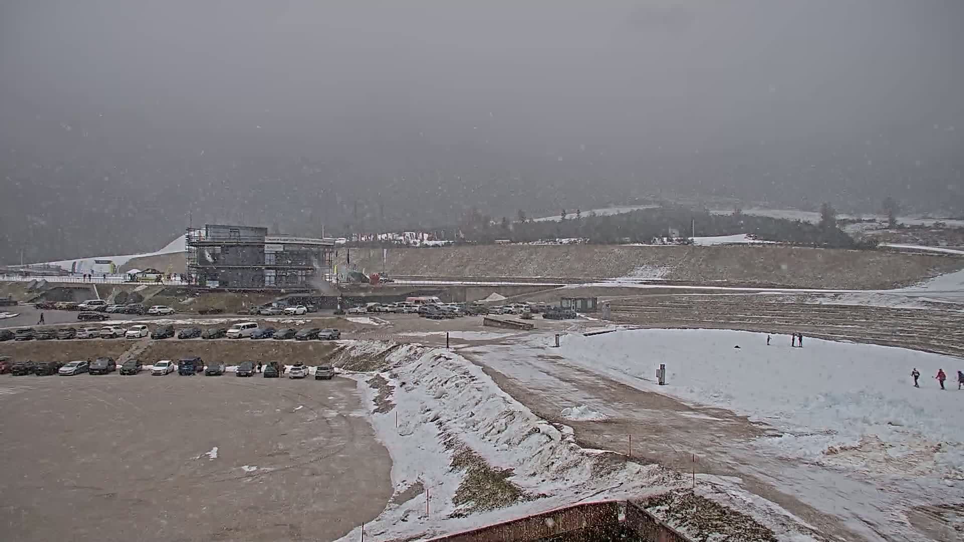A large parking lot filled with cars is situated near a modern building in a snowy, overcast landscape.