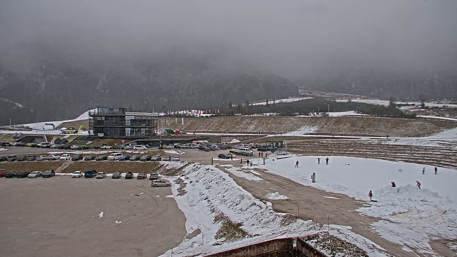 A snowy mountainside parking lot with a multi-story building under construction, a partially snow-covered field with a few people, and a foggy, overcast sky.