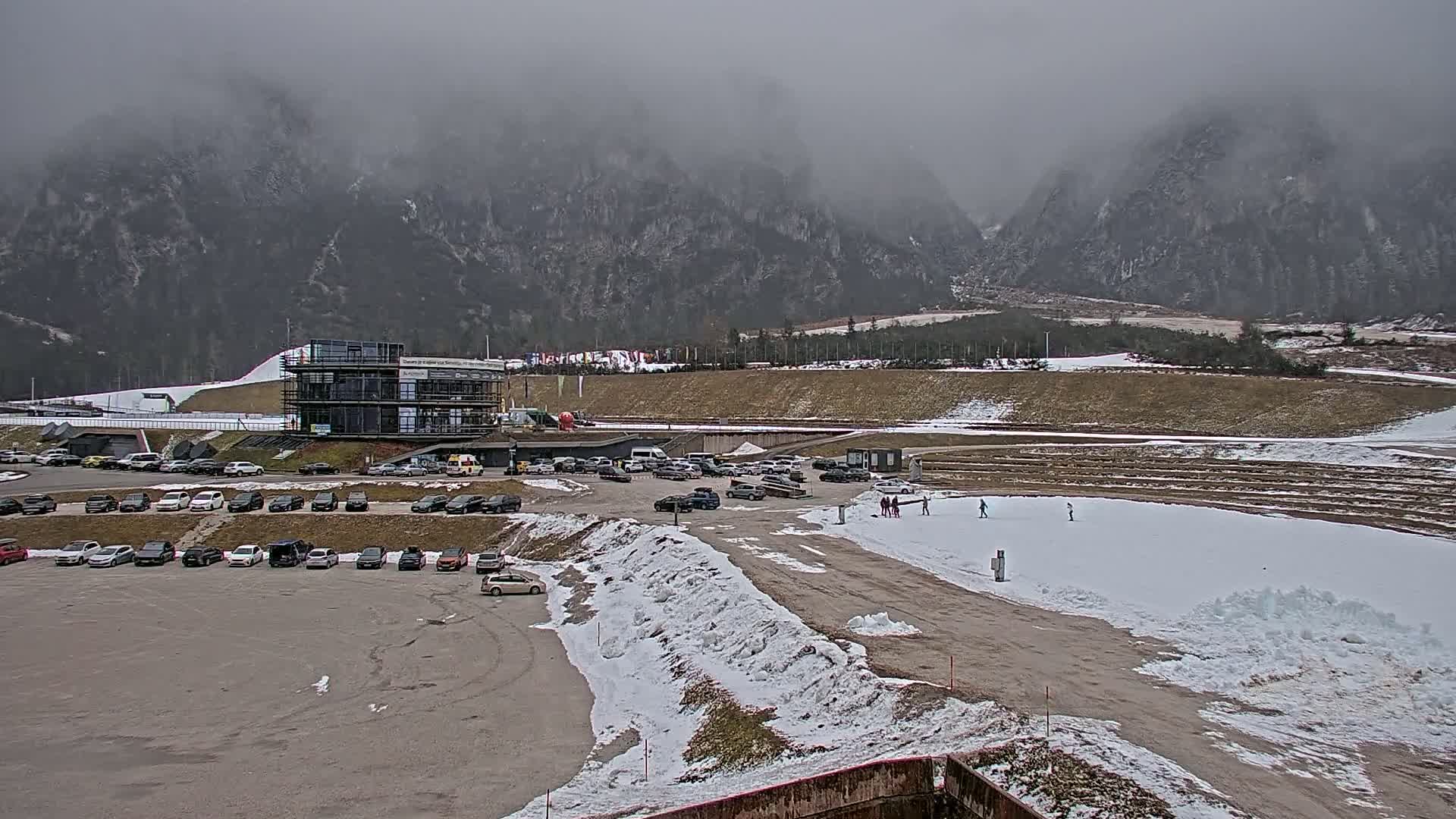 A parking lot filled with cars sits in front of a modern building, with a snowy, flat area and several people visible in the distance, all set against a backdrop of fog-covered mountains.