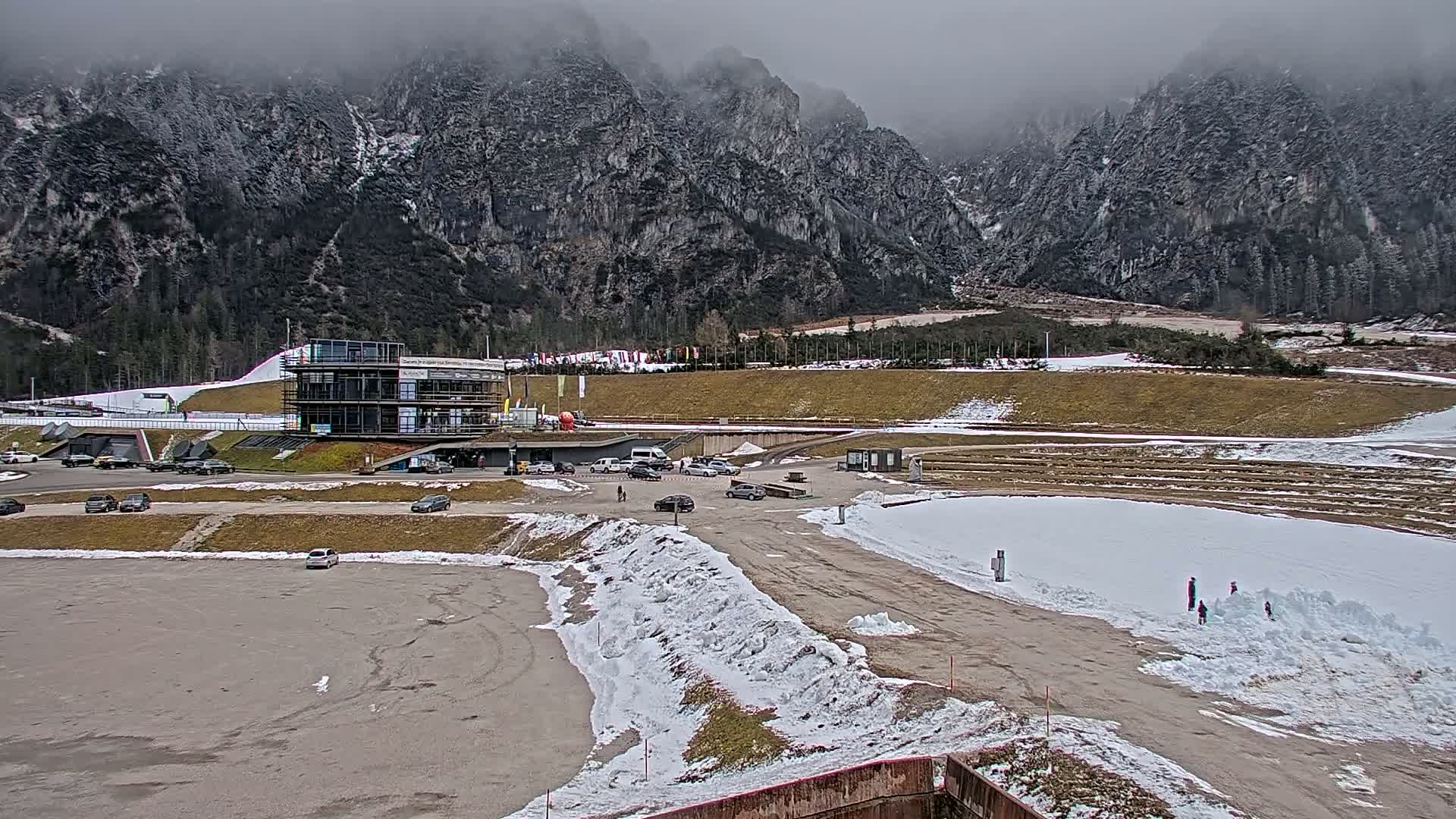 A modern building overlooks a snow-dusted parking lot and a snowy area with several people playing in the snow, all set against a backdrop of large, fog-covered mountains.