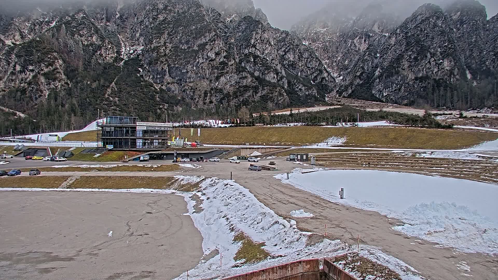 A modern building sits at the base of snow-dusted mountains on an overcast day, with a parking lot and patches of snow in the foreground.