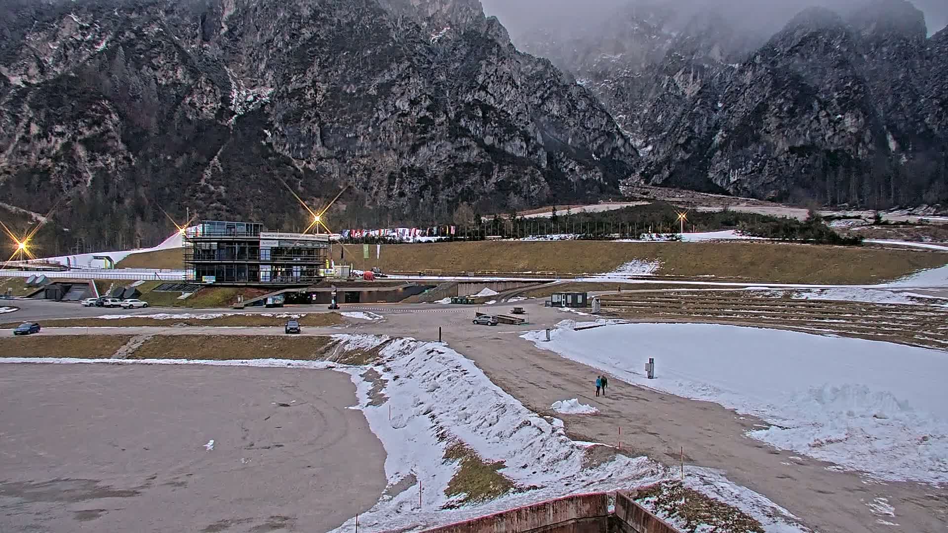 A snowy outdoor scene shows a modern building near a mountain range under an overcast sky, with a few cars parked nearby and two people walking on a path.