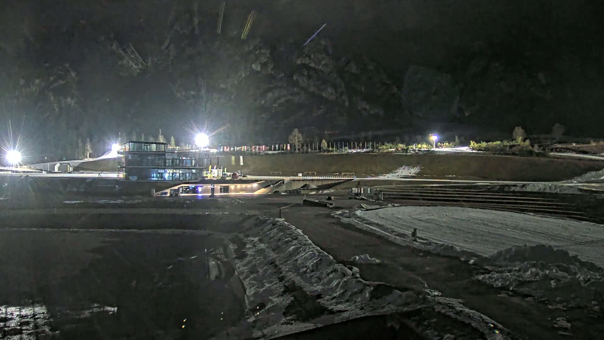 A nighttime view of an outdoor sports facility, partially snow-covered, with floodlights illuminating a modern building and surrounding grounds.