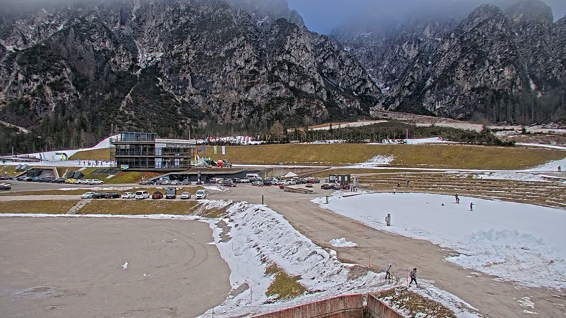 A parking lot with cars is situated next to a multi-story building near a snowy, mountainous area where several people are seen skiing and walking.