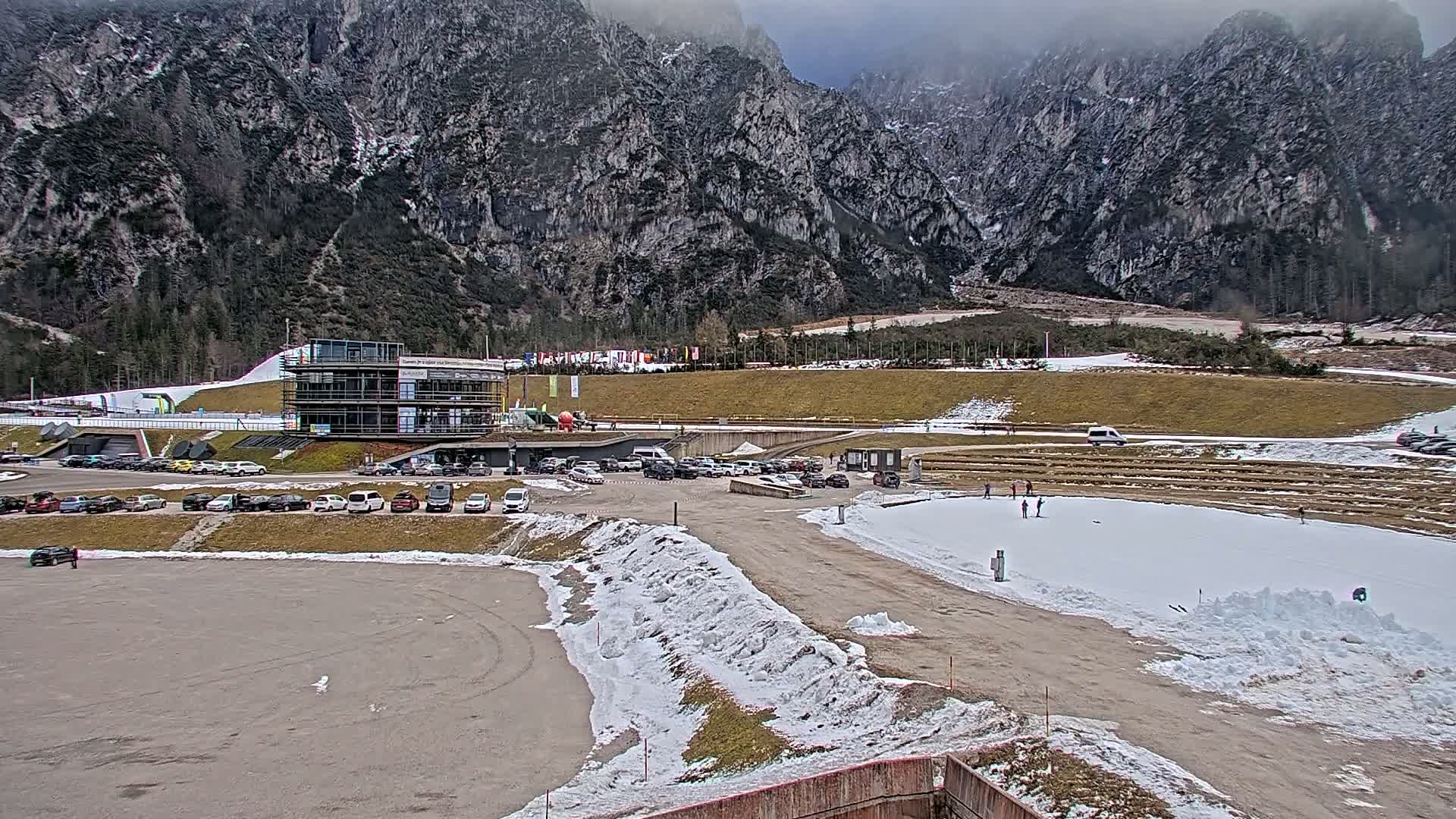 A parking lot filled with cars sits below a multi-story building next to a snow-covered area in a valley surrounded by large, grey mountains under an overcast sky.