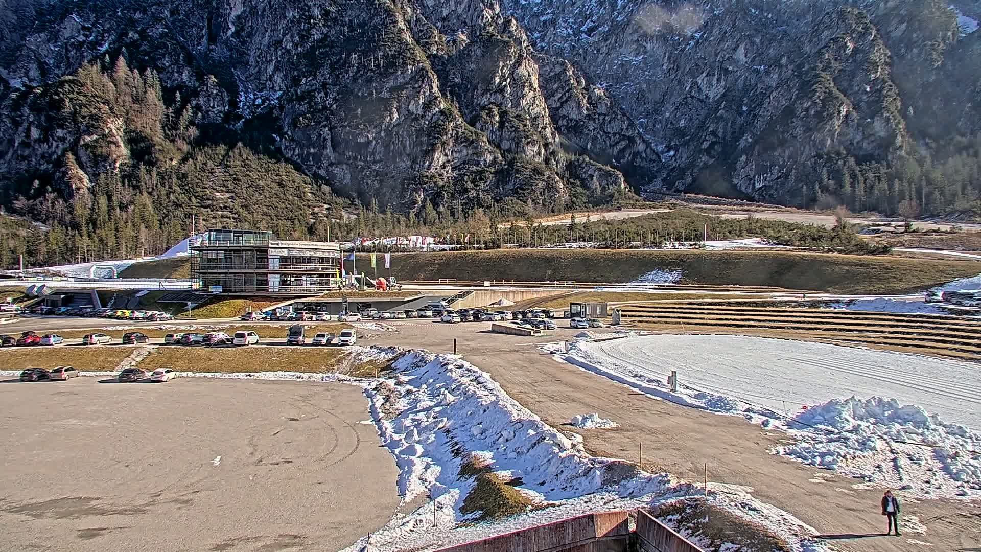 A parking lot with cars is situated in front of a multi-story building, next to a snow-covered track and a mountainous backdrop on a sunny day with patches of snow on the ground.