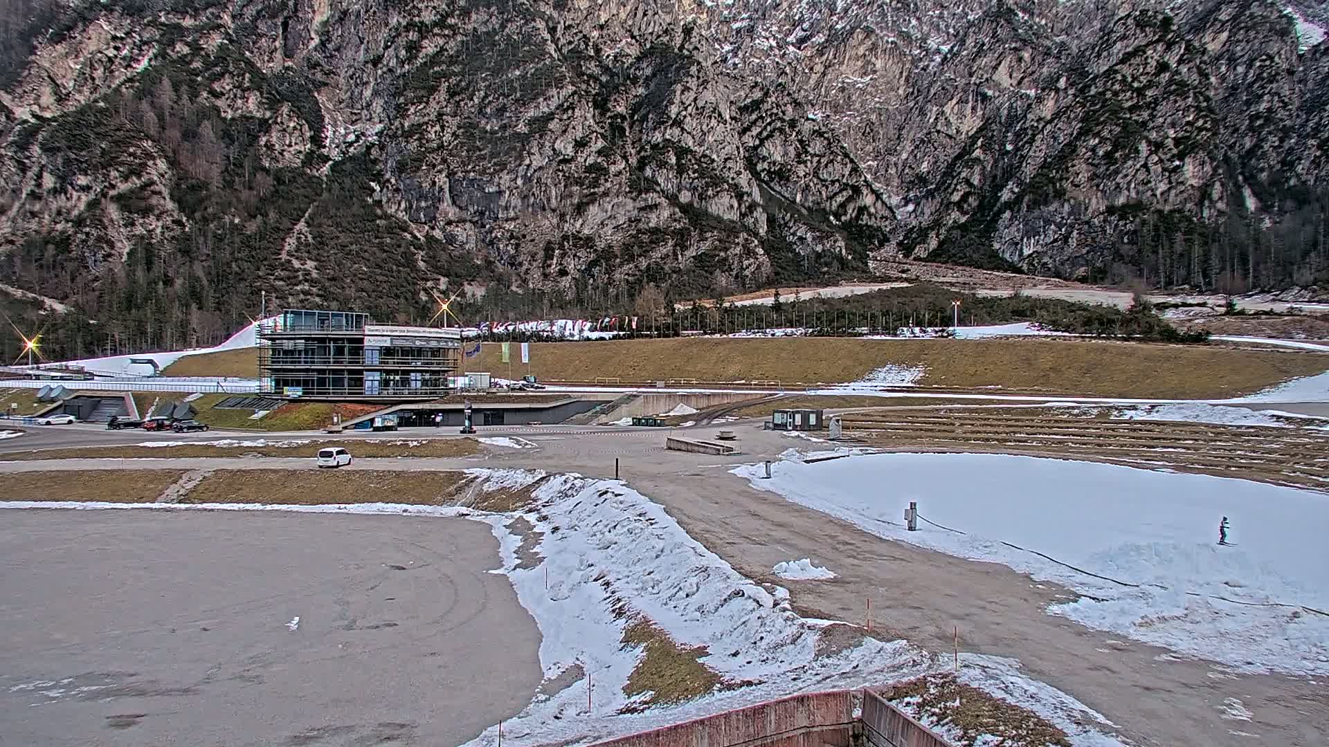 A multi-story building sits at the base of a large, rocky mountain, with a snowy, paved area in the foreground, and patches of snow scattered throughout the scene.