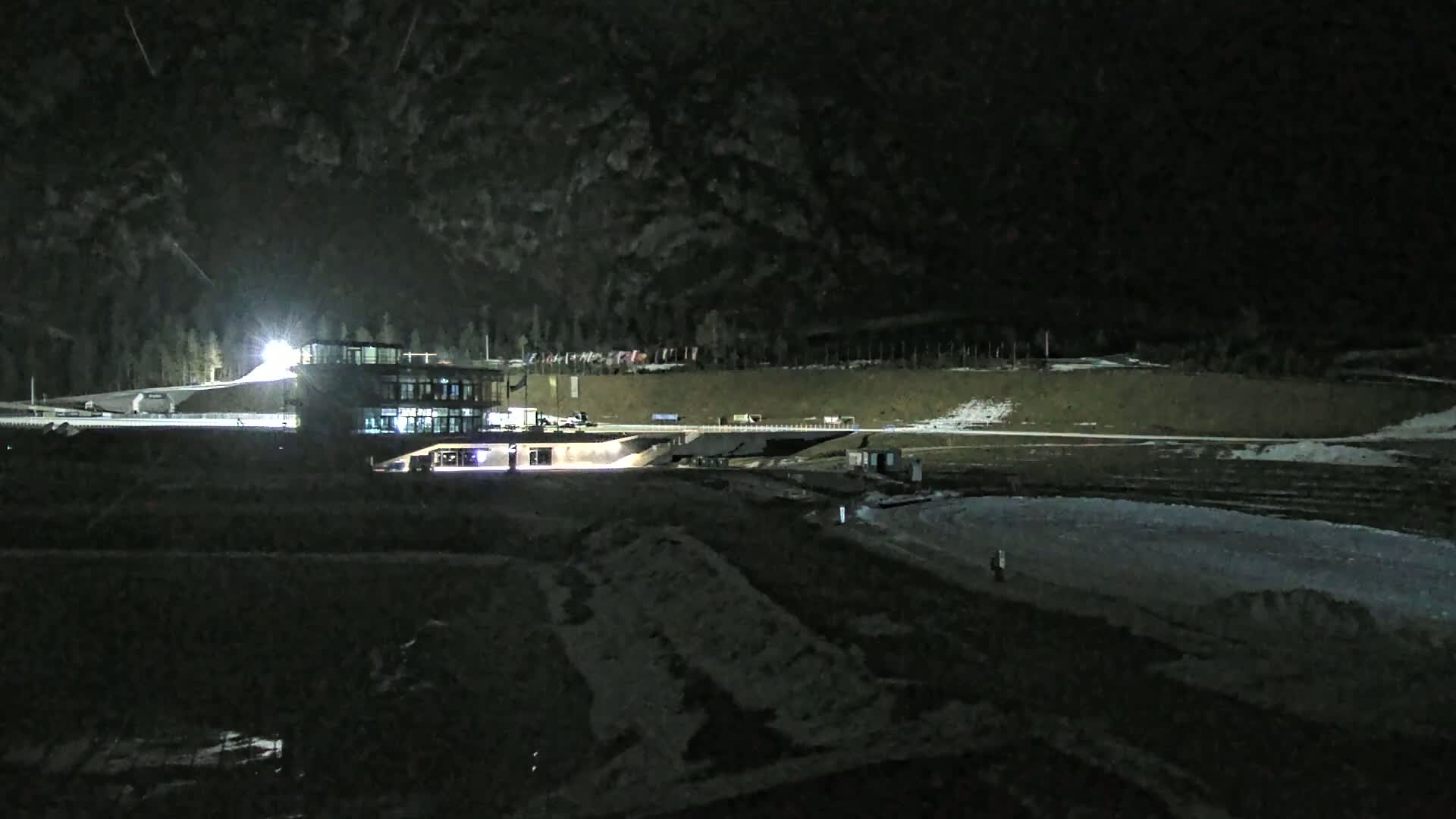 A nighttime, snow-dusted outdoor scene shows a lit building near a large, dark, empty area, possibly a frozen pond or rink.