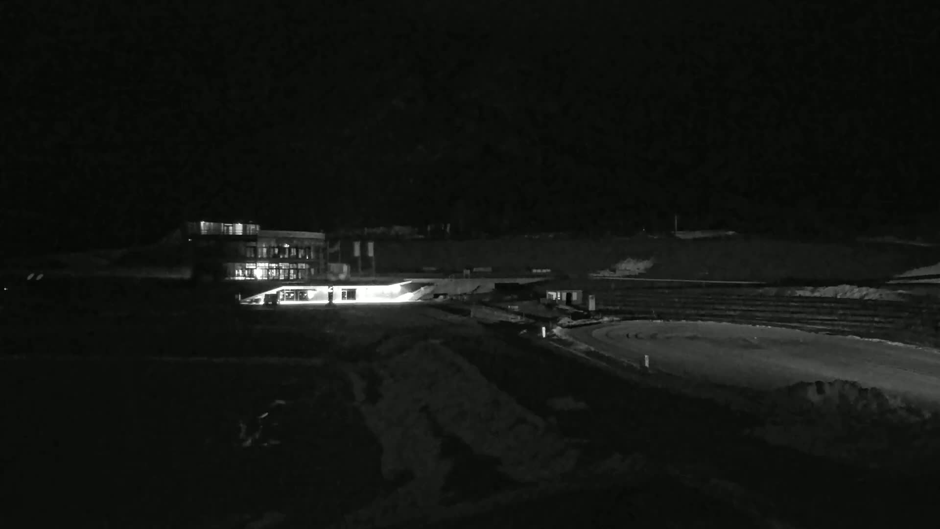 A brightly lit building sits near a dark, snow-covered oval track at night.