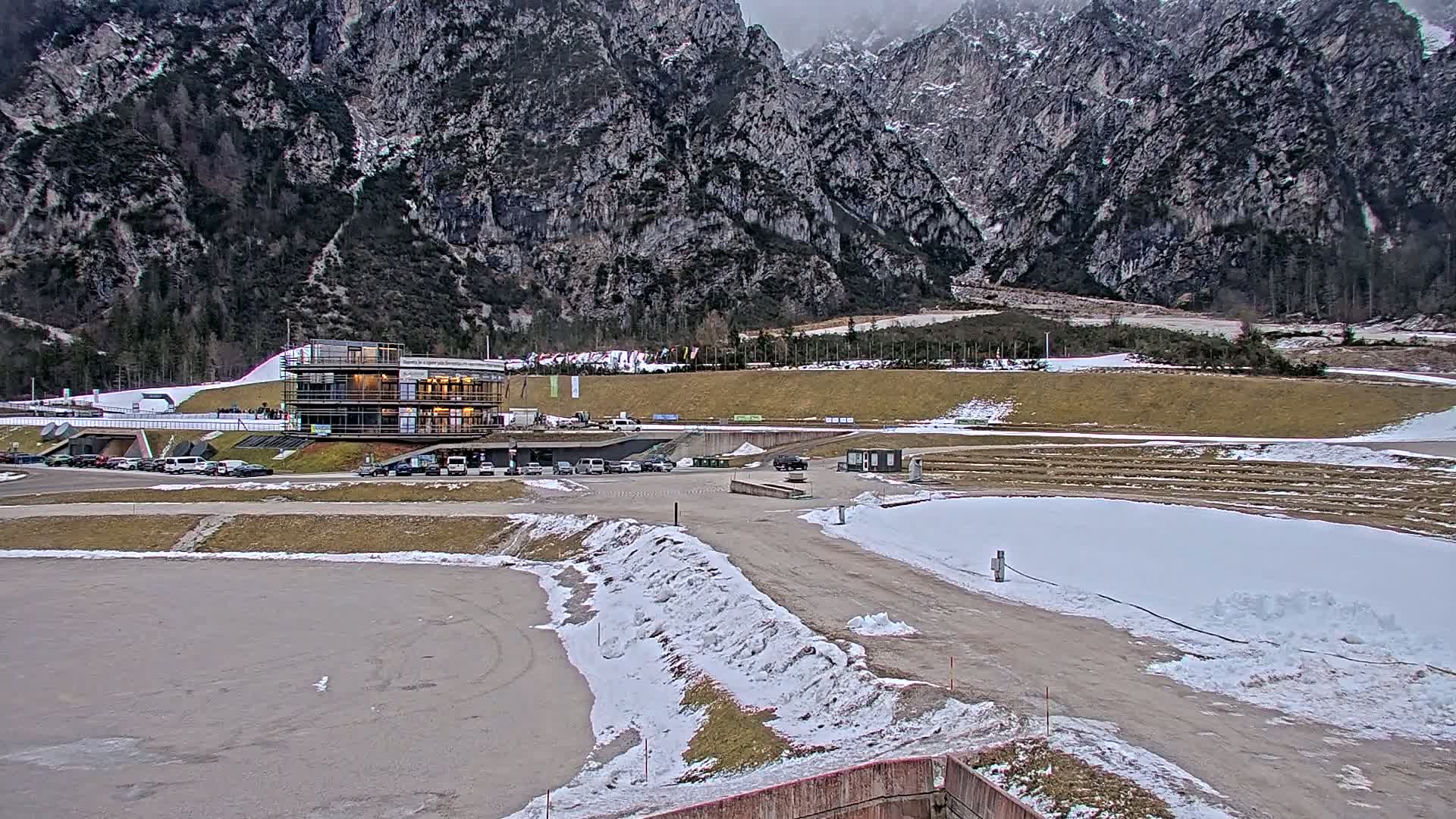 A parking lot with cars in front of a multi-story building sits at the base of snow-dusted mountains under an overcast sky.