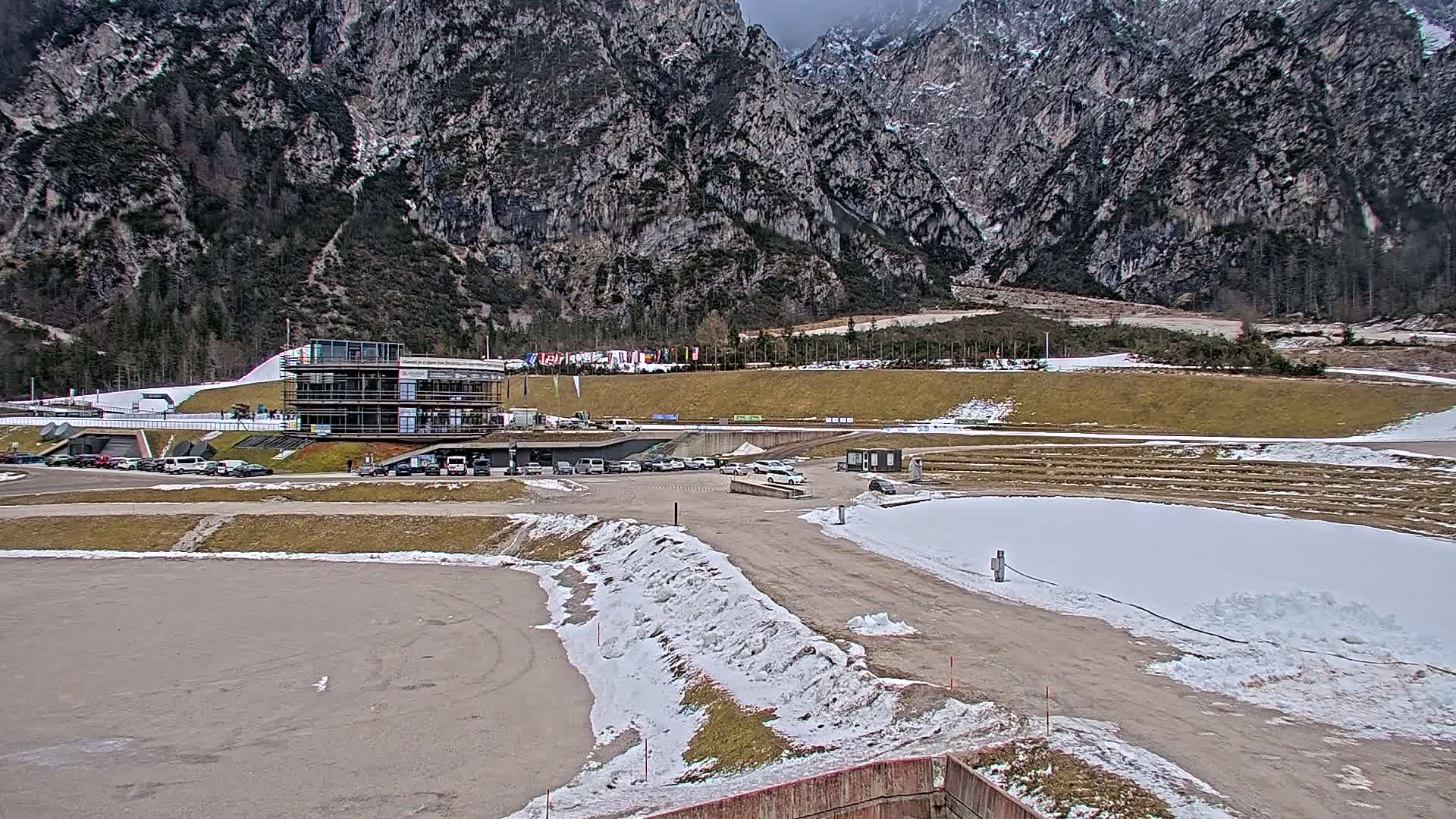 A parking lot and a multi-story building sit at the base of a snow-dusted mountain range on an overcast day.