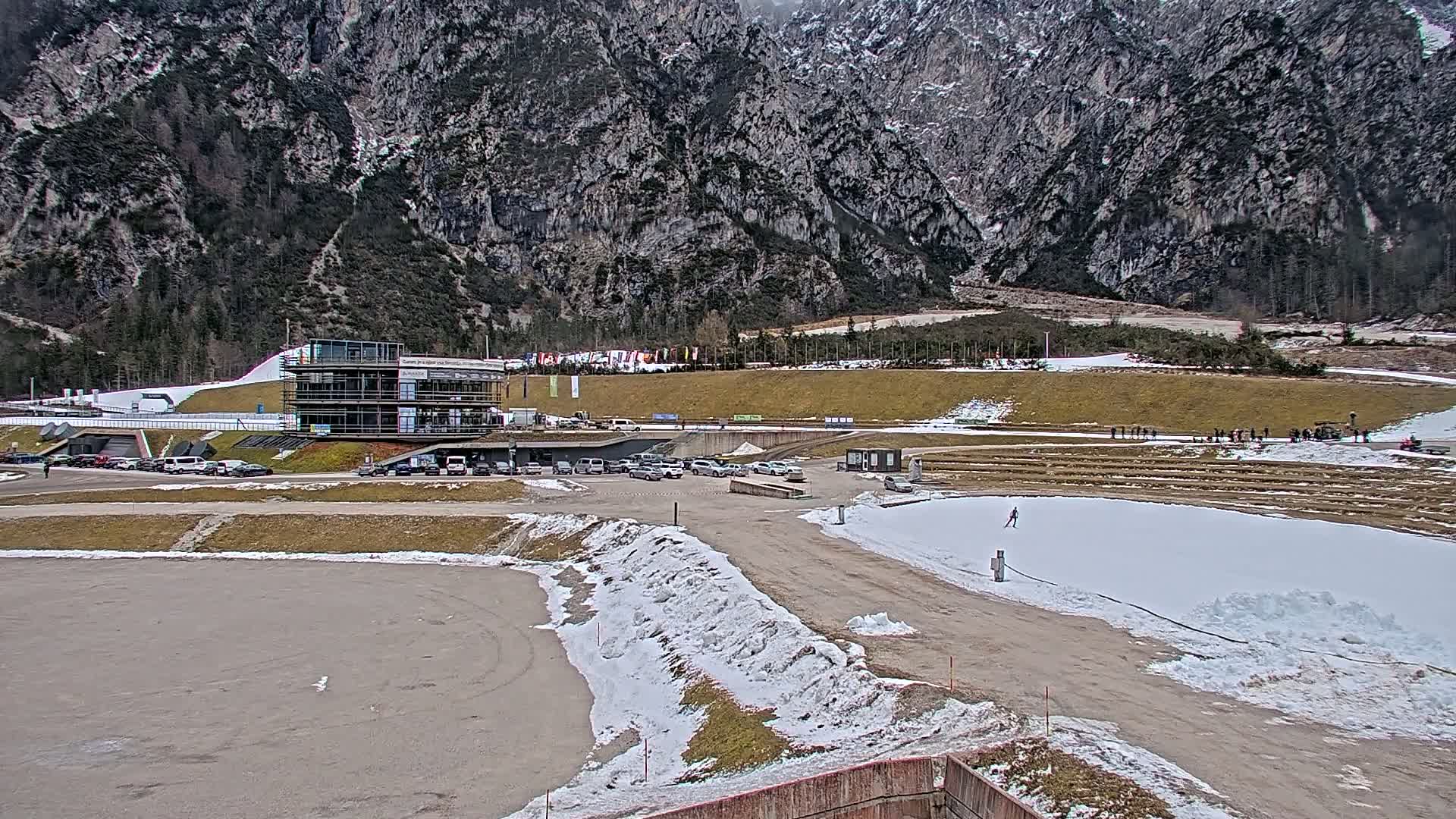 A modern building sits in a parking lot near a snowy biathlon range set against a backdrop of large, gray mountains on an overcast day.