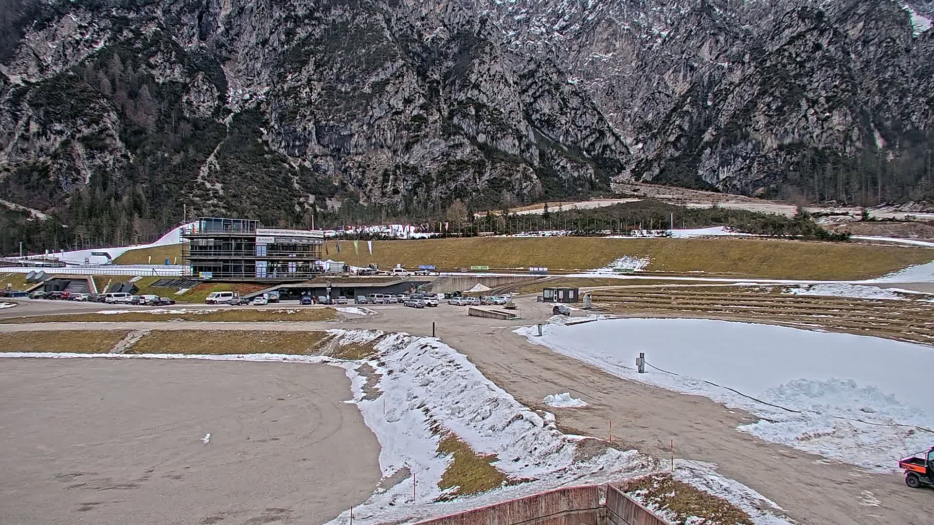 A multi-story building sits in a parking lot next to a snow-covered, outdoor athletic facility in a mountainous area with patchy snow on the ground.