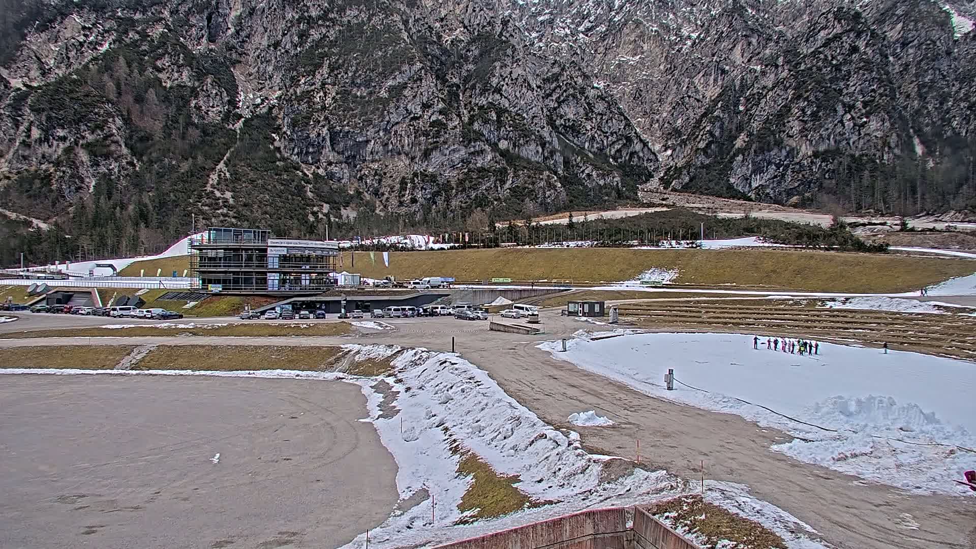 A multi-story building sits in a parking lot near a snowy, outdoor athletic facility at the base of a large, rocky mountain.