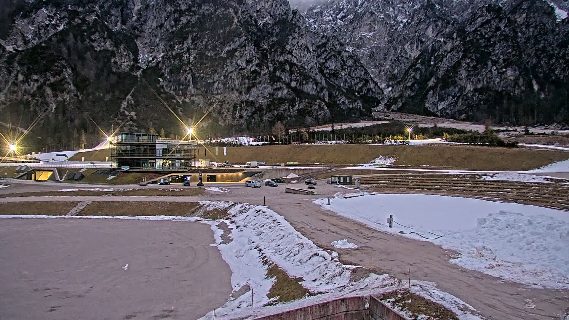 A modern building sits in a snowy, mountainous landscape at dusk, with several vehicles parked nearby.