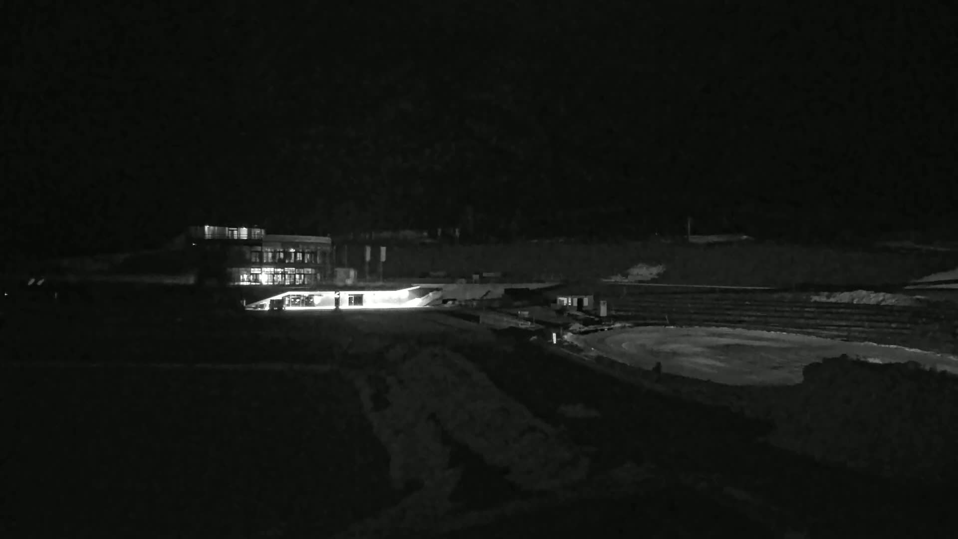 A brightly lit building is visible at night, near a dark, snow-covered oval-shaped area.