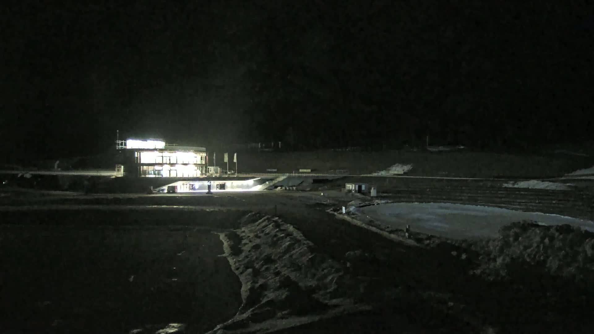 At night, a brightly lit building stands near a dark, snow-covered outdoor area that includes a frozen body of water.