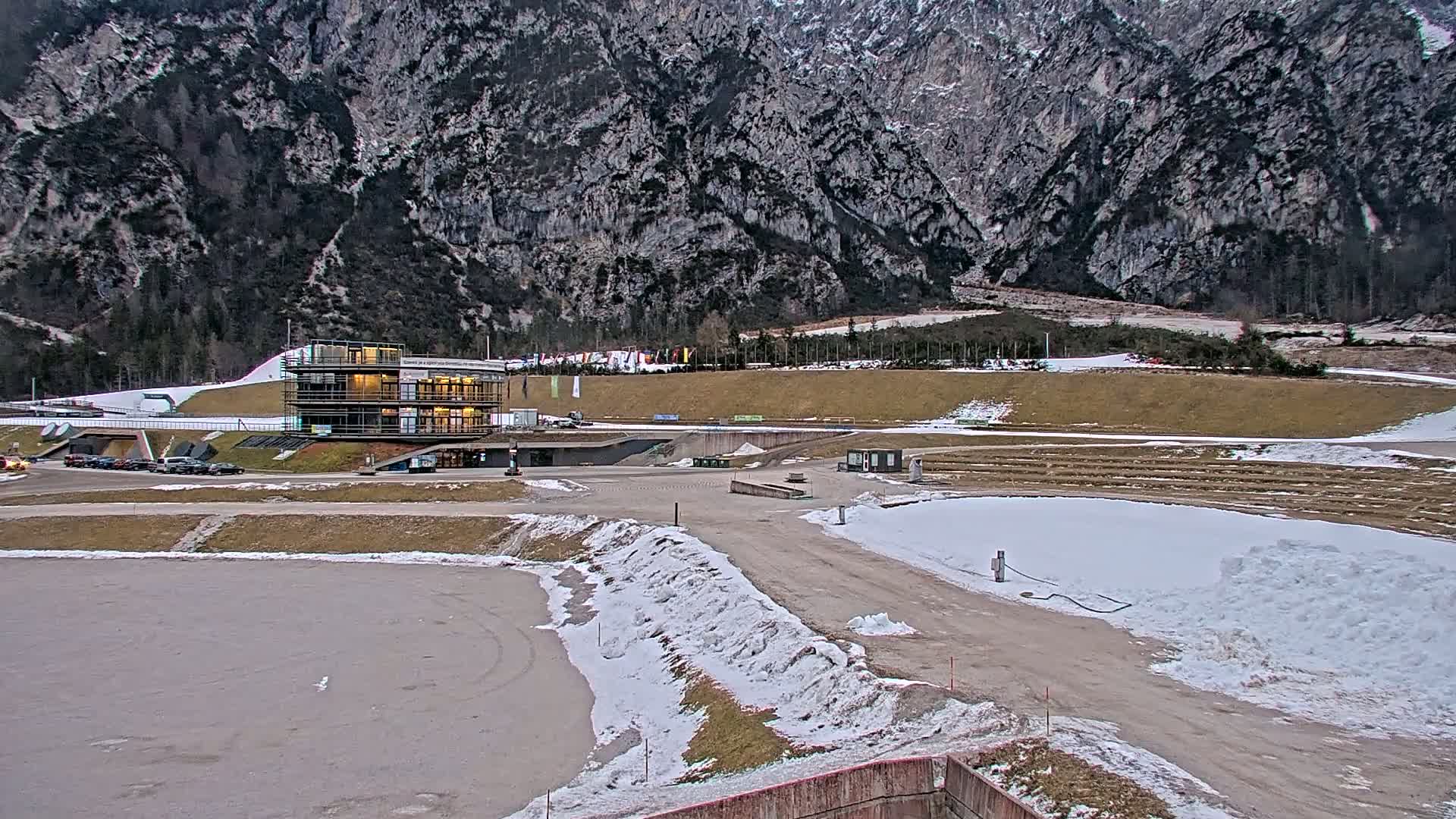 A partially snow-covered outdoor sports complex, featuring a modern building, sits at the base of a large, rocky mountain under a cloudy sky.