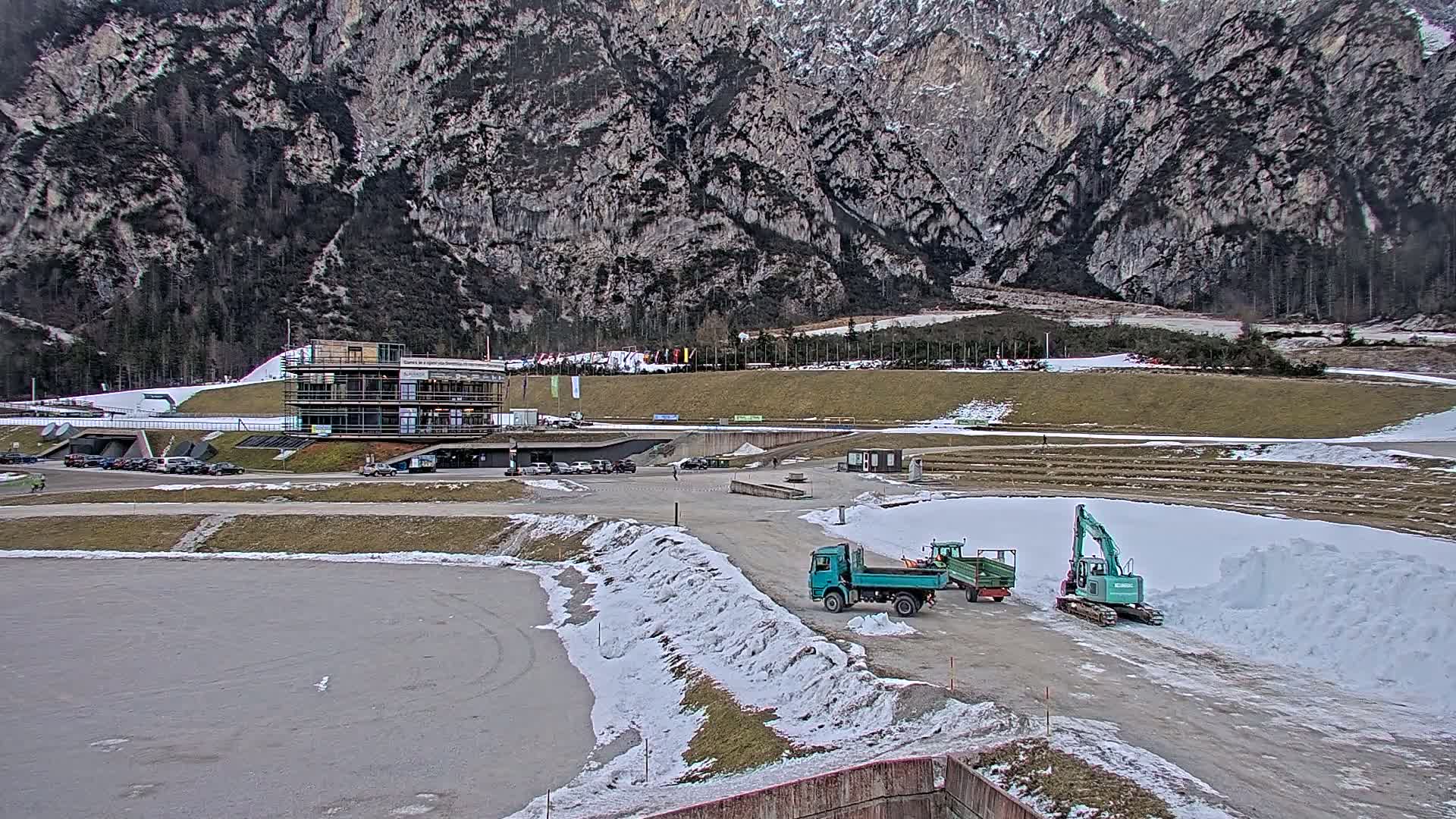 A partially snow-covered outdoor area with a building, several vehicles including a dump truck and excavator, and a large mountain in the background.