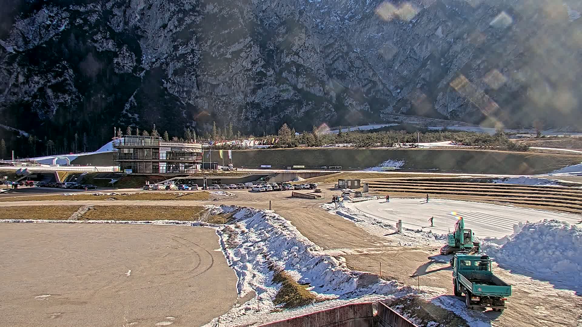 A sunny day reveals a snowy outdoor sports complex nestled against a large mountain, with construction equipment present.