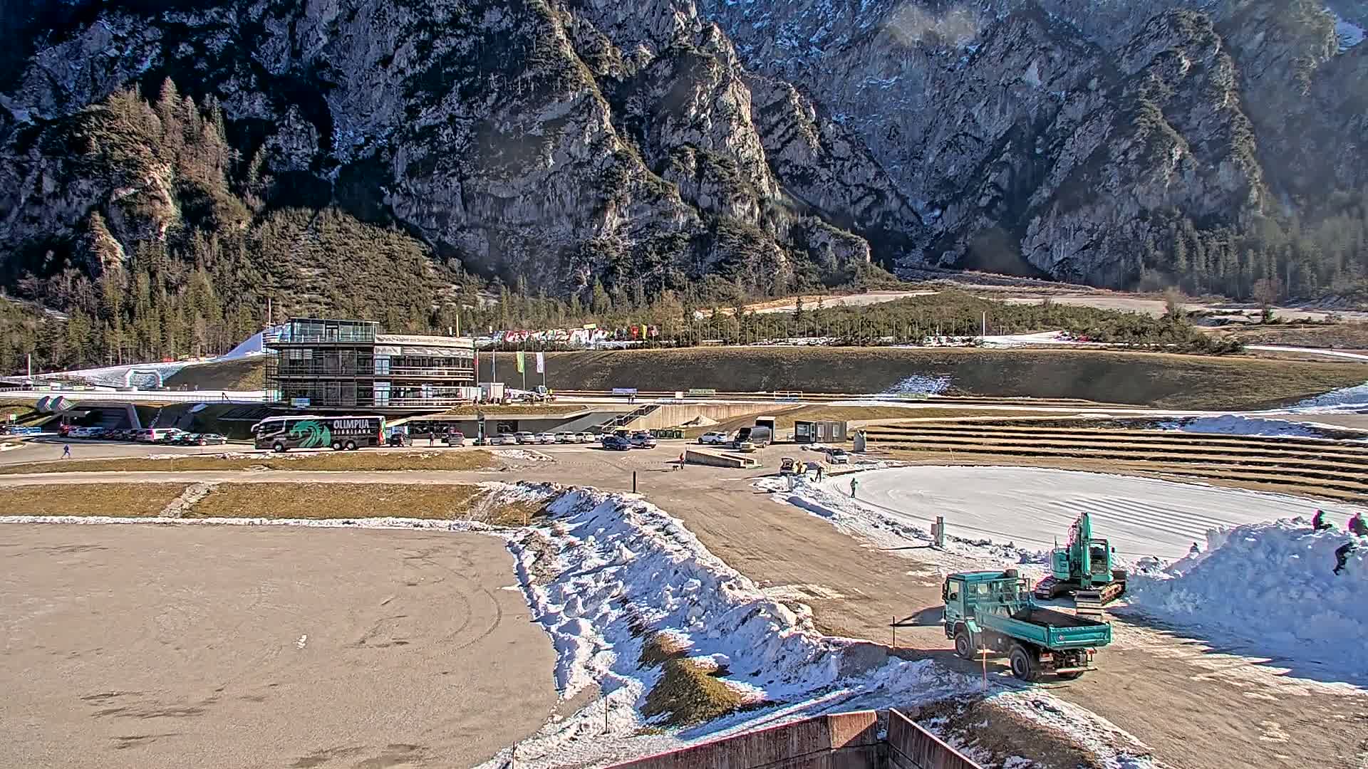 A sunny day at a snow-covered outdoor sporting venue nestled in a mountainous region, featuring a multi-story building, vehicles, and snow-removal equipment.