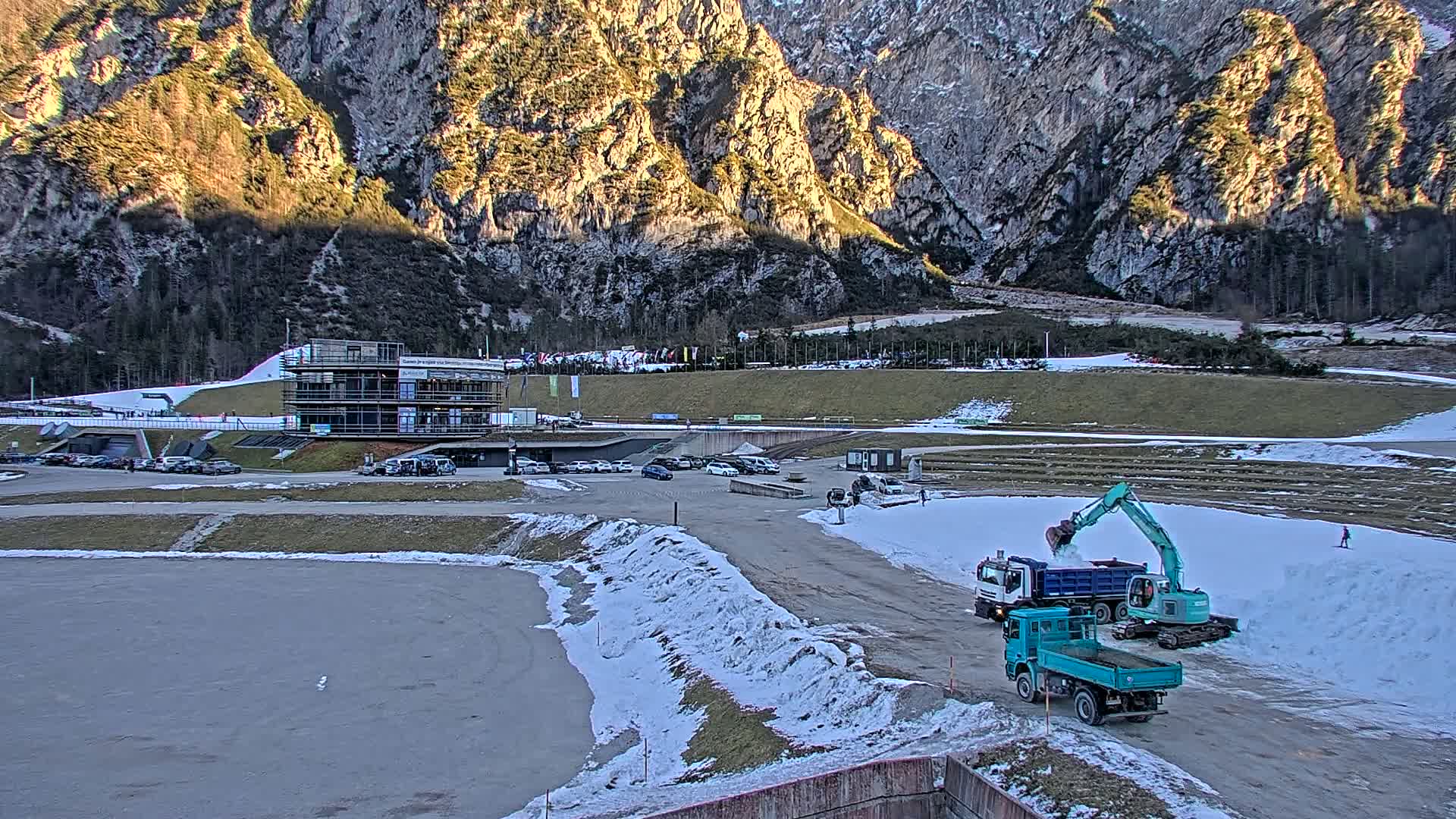 A teal excavator is moving snow into a dump truck in a parking lot next to a multi-story building at the base of a large, rocky mountain under partly sunny conditions.