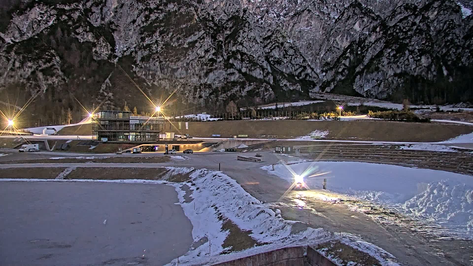 A modern building complex is illuminated at night, situated in a snowy area at the base of a large mountain range under dark skies.