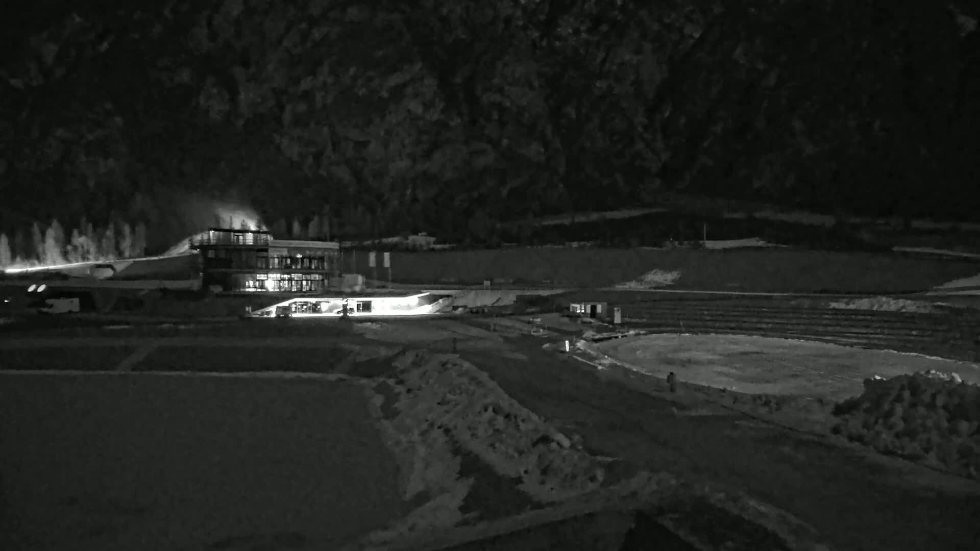 A nighttime, snow-covered landscape features a lit building near a flat, possibly icy, oval area, with snow blowing or spraying from a structure nearby.