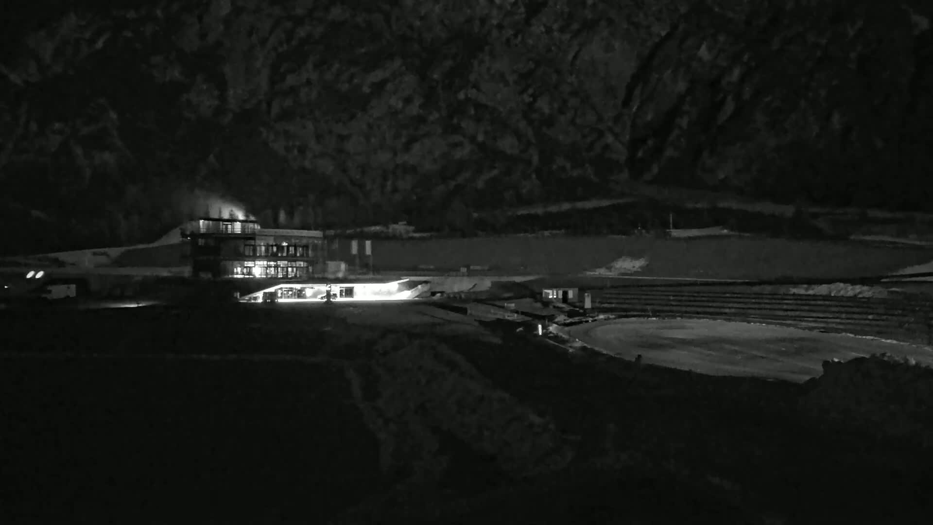 A nighttime, low-light view of a lit building near a dark oval-shaped area, possibly a track or rink,  with a mountainous background.