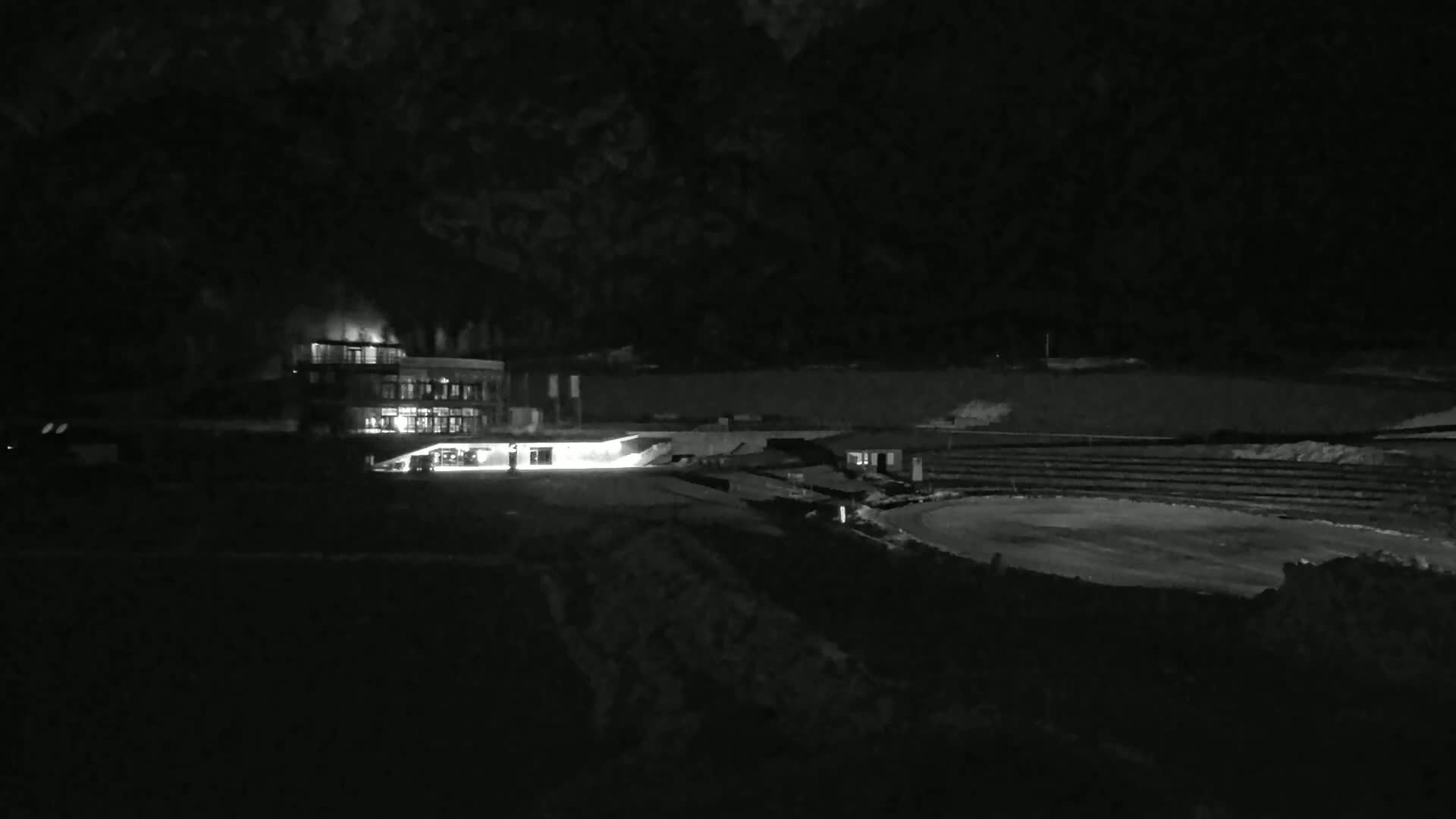 A brightly lit building is visible at night, along with a dark, empty oval-shaped structure and surrounding land.