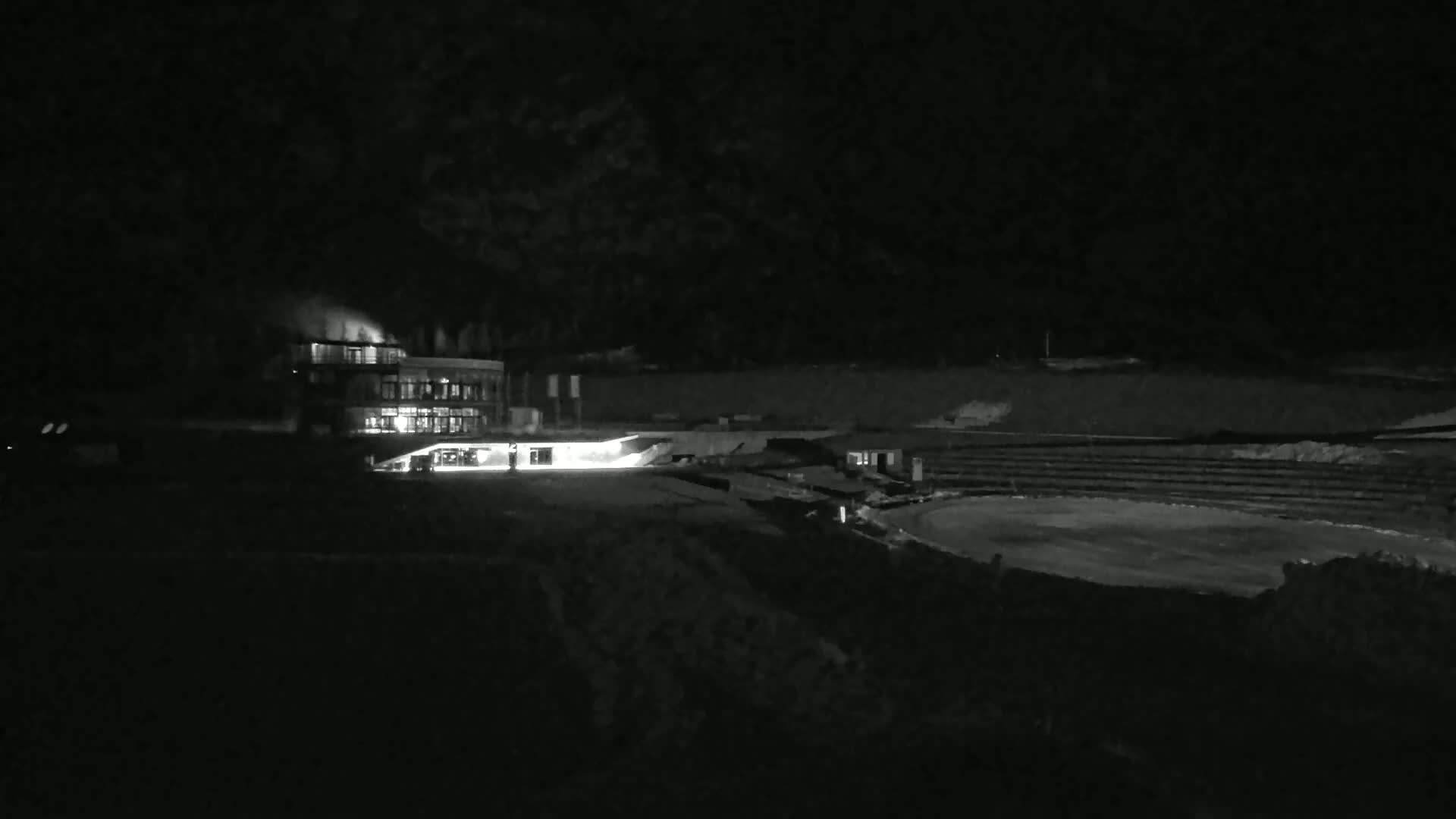 A brightly lit building sits near a dark, seemingly empty amphitheater-like structure at night.