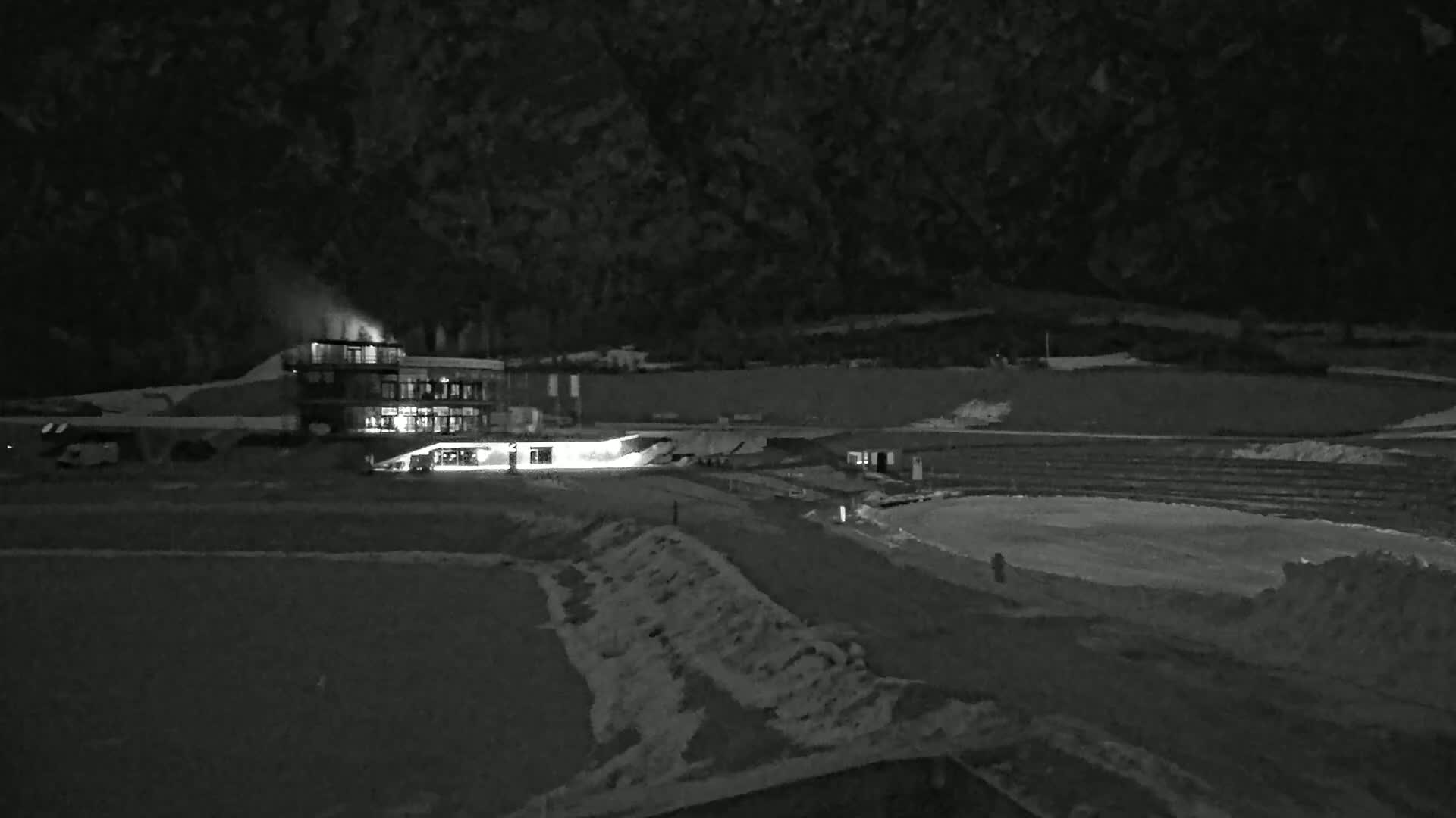 A nighttime, low-light image shows a building illuminated with several lights, near a seemingly snow-covered, flat area with piles of dirt or snow, all set against a dark, mountainous backdrop.