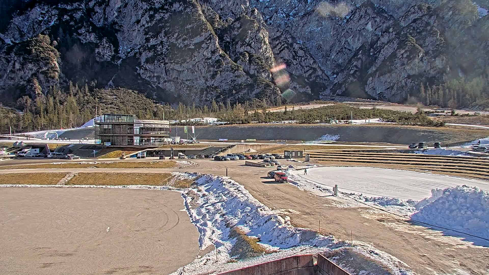 A modern building sits near a paved area with several cars parked, next to an oval-shaped ice rink partially covered in snow, all set against a backdrop of a mountainous landscape on a sunny day.