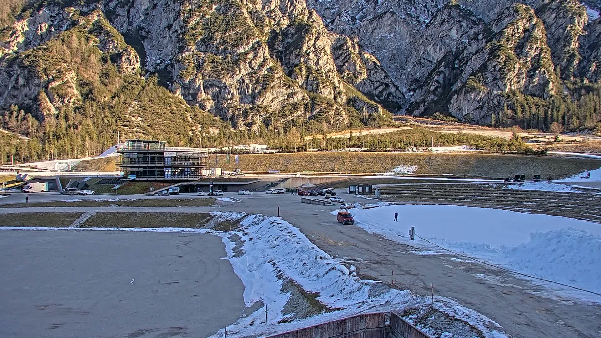 A modern building sits in a parking lot next to a snowy, mountainous landscape, with several vehicles parked nearby under a sunny sky.