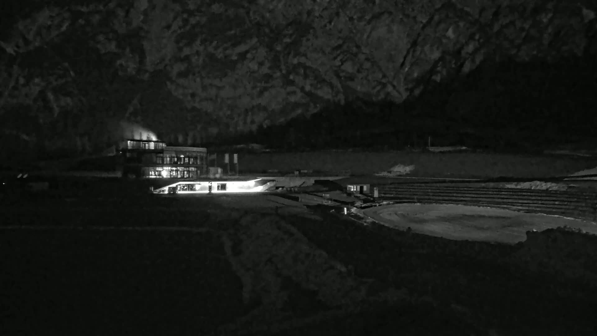 A brightly lit building sits near a dark, snow-covered oval structure at night, against a backdrop of dark, mountainous terrain.