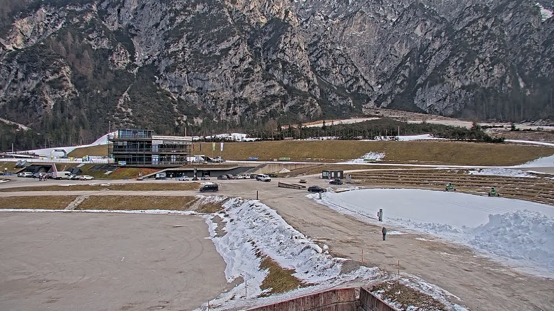 A modern building sits in a parking lot near a snowy track, with a large mountain in the background under a cloudy sky.