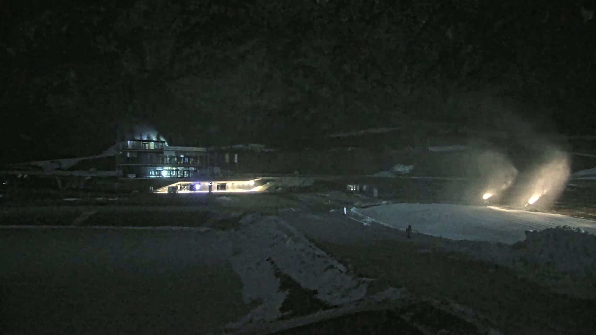 At night, snow cannons are operating on a snow-covered area in front of a lit building.