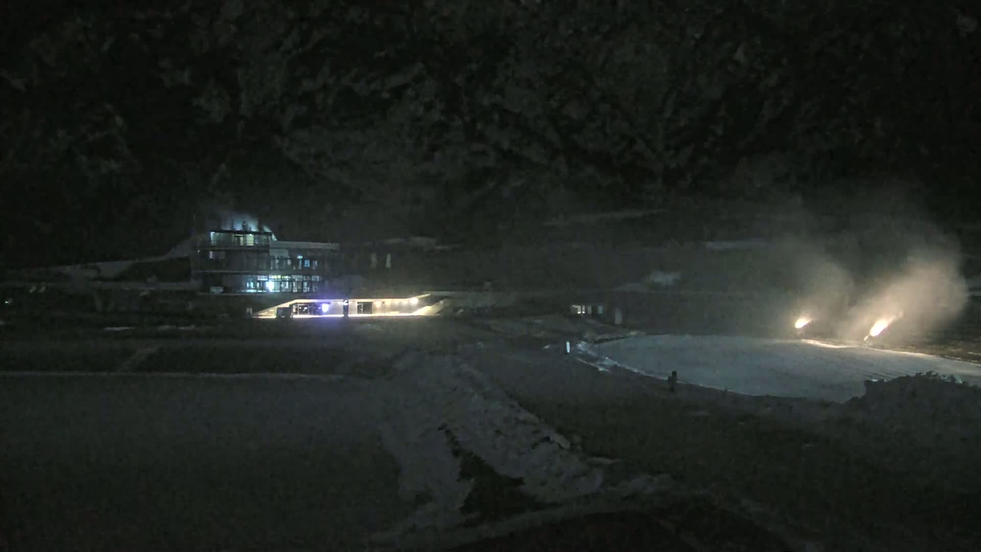 At night, two snow cannons emit plumes of snow onto a snow-covered area in front of a lit building.