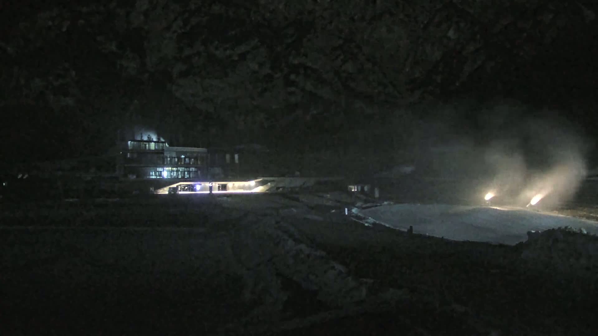 At night, a lit building is situated near a flat, light-colored surface where two bright lights are emitting smoke.